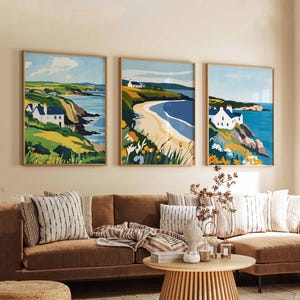 May include: Three framed landscape art prints depicting coastal scenes with white houses, green hills, and blue water. The artwork has a modern, minimalist style with a focus on color and shape. The frames are a light wood color.