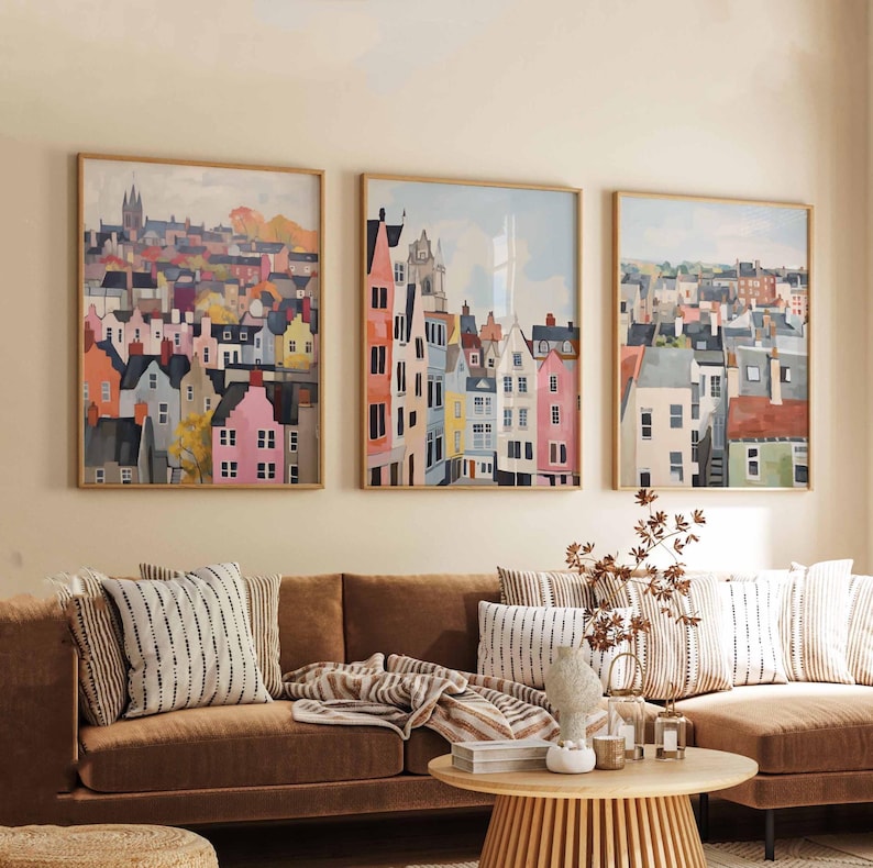 May include: Three framed watercolor paintings of colorful European townhouses hang above a brown sofa with striped pillows. The artwork features buildings in shades of pink, orange, blue, and yellow, with wooden frames.