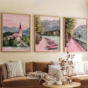 May include: Three framed landscape art prints. The prints depict a lake scene with mountains, trees, and buildings. The colour palette includes pink, green, and brown tones. The artwork is displayed above a brown sofa with striped cushions.