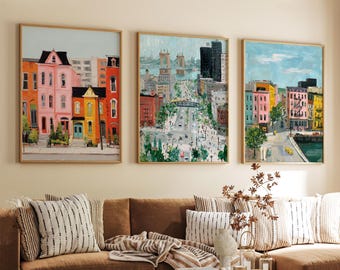 May include: Three framed cityscape paintings. The artwork features colorful buildings and street scenes. The paintings are in wooden frames and are displayed above a brown sofa with striped pillows. The art adds a touch of urban charm to the living space.