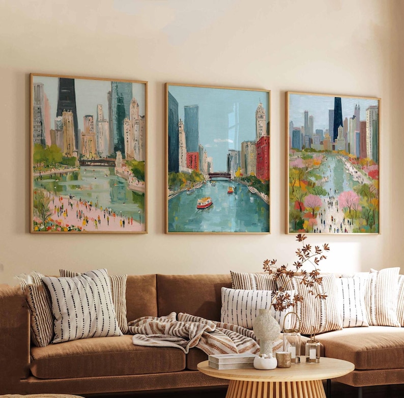 May include: Three framed art prints depicting a cityscape with a river and buildings. The paintings feature a palette of blues, greens, and pinks, with people and boats visible. The frames are a light wood color, and the artwork is displayed above a brown sofa.