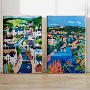 May include: Two framed landscape paintings. The left painting depicts a town with a river, white buildings, and green hills. The right painting shows a similar town with a river, orange and green foliage, and a blue sky.
