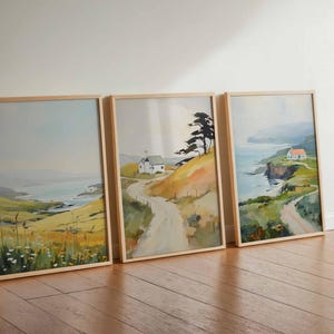 May include: Three framed landscape paintings depicting coastal scenes with rolling hills, winding roads, and small houses. The artwork features a muted color palette of blues, greens, and yellows, with wooden frames. The paintings are leaning against a white wall on a wooden floor.