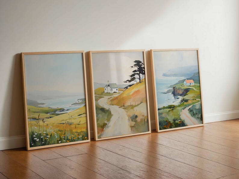 May include: Three framed landscape paintings depicting coastal scenes with rolling hills, winding roads, and small houses. The artwork features a muted color palette of blues, greens, and yellows, with wooden frames. The paintings are leaning against a white wall on a wooden floor.