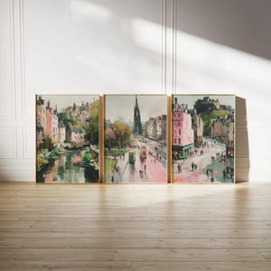 May include: Set of three framed watercolor prints depicting a city scene with buildings, a river, and a street with people. The artwork features a muted color palette with pink, green, and brown tones. The frames are gold.