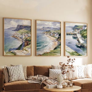 Può includere: Tre dipinti ad acquerello incorniciati di scene costiere. L'opera raffigura una cittadina balneare con un porto, una spiaggia e delle scogliere. I dipinti sono in cornici di legno e sono esposti sopra un divano marrone.