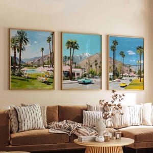 May include: Three framed landscape paintings depicting a desert scene with palm trees, mountains, and vintage cars. The artwork features a color palette of blues, greens, and pinks, with a mid-century modern aesthetic. The paintings are displayed above a brown sofa with decorative pillows.