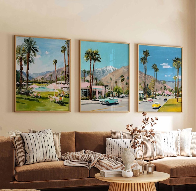 May include: Three framed landscape paintings depicting a desert scene with palm trees, mountains, and vintage cars. The artwork features a color palette of blues, greens, and pinks, with a mid-century modern aesthetic. The paintings are displayed above a brown sofa with decorative pillows.