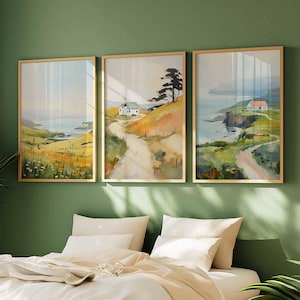 May include: Three framed landscape art prints. Each print features a coastal scene with a road, a house, and a body of water. The frames are gold-colored, and the artwork has a watercolor style with muted colors.