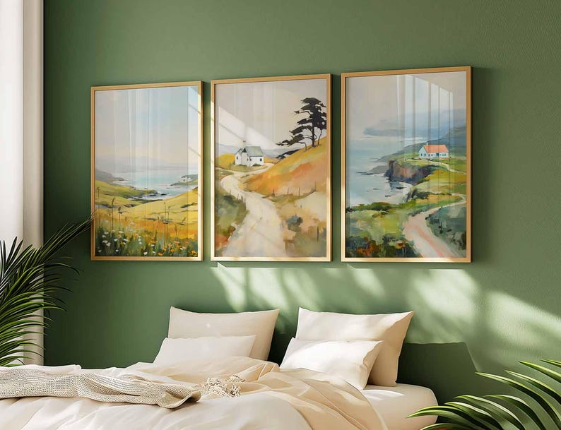 May include: Three framed landscape art prints. Each print features a coastal scene with a road, a house, and a body of water. The frames are gold-colored, and the artwork has a watercolor style with muted colors.