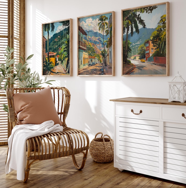 May include: Three framed paintings of tropical scenes with palm trees, colorful buildings, and lush greenery. The paintings are hung on a white wall above a white cabinet with a wooden top.