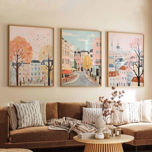 May include: Three framed art prints depicting colorful cityscapes with buildings, trees, and streets. The prints are hung on a light beige wall above a brown couch with striped pillows and a throw blanket.