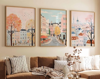 May include: Three framed art prints depicting colorful cityscapes with buildings, trees, and streets. The prints are hung on a light beige wall above a brown couch with striped pillows and a throw blanket.