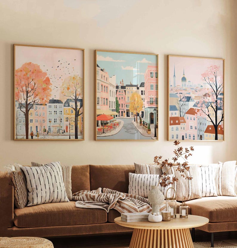 May include: Three framed art prints depicting colorful cityscapes with buildings, trees, and streets. The prints are hung on a light beige wall above a brown couch with striped pillows and a throw blanket.