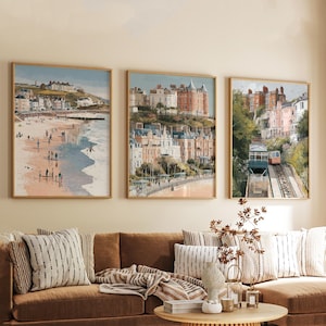 May include: Three framed watercolour prints of coastal scenes. The first shows a beach with people, the second a town on a hill, and the third a funicular railway. The prints have wooden frames and are displayed above a brown sofa with cushions.