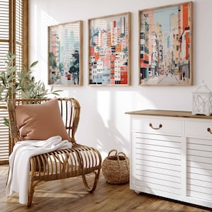 May include: A wicker chair with a white throw blanket and a peach colored pillow. The chair is in front of a white wall with three framed pictures of cityscapes. The pictures are in shades of pink, blue, and green. A white cabinet with a wood top is to the right of the chair. A wicker basket is in front of the cabinet.