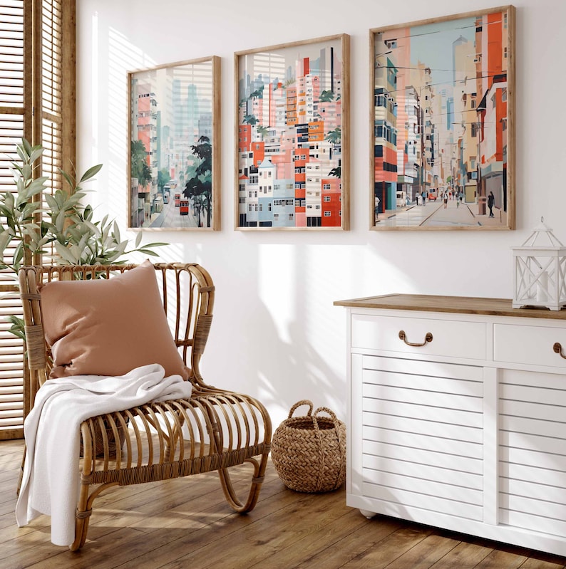May include: A wicker chair with a white throw blanket and a peach colored pillow. The chair is in front of a white wall with three framed pictures of cityscapes. The pictures are in shades of pink, blue, and green. A white cabinet with a wood top is to the right of the chair. A wicker basket is in front of the cabinet.