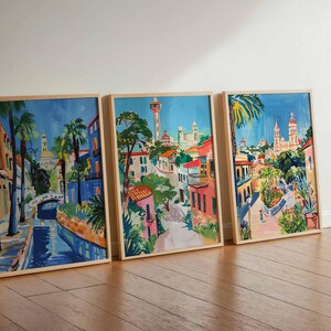 May include: Three framed watercolor paintings depicting a colorful cityscape with buildings, palm trees, and a blue waterway. The paintings are in wooden frames and are hung on a white wall.
