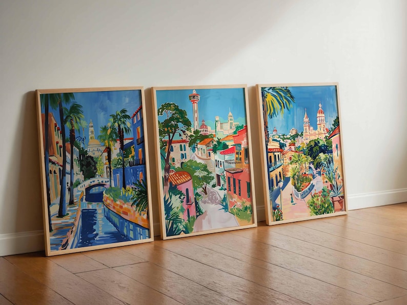 May include: Three framed watercolor paintings depicting a colorful cityscape with buildings, palm trees, and a blue waterway. The paintings are in wooden frames and are hung on a white wall.