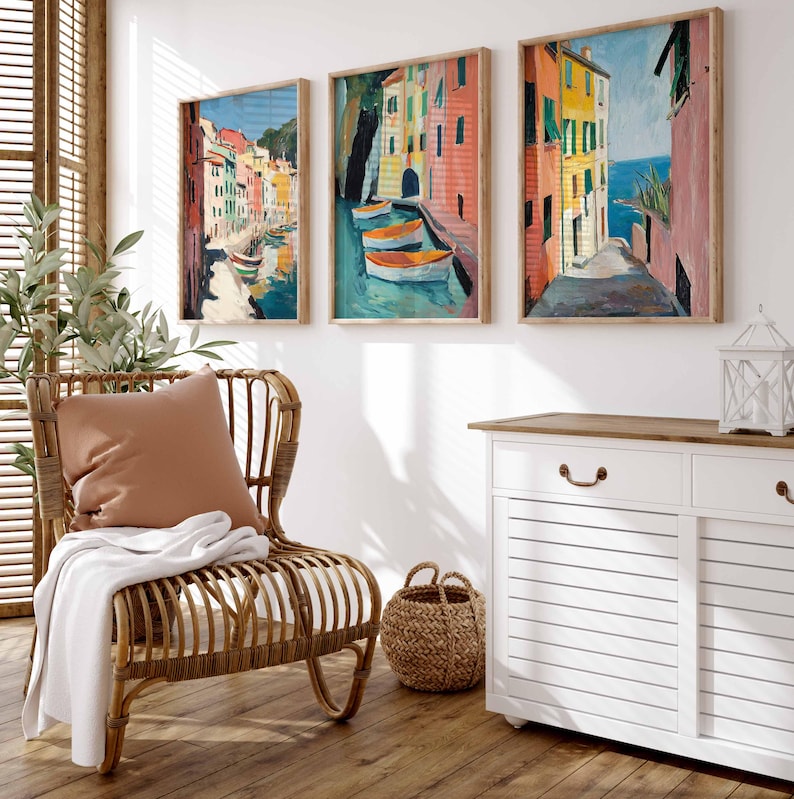 May include: Three framed paintings of colorful Italian buildings, one with boats in a canal, another with a street scene, and the third with a view of the ocean. The paintings are hanging on a white wall above a white dresser with a wooden top.