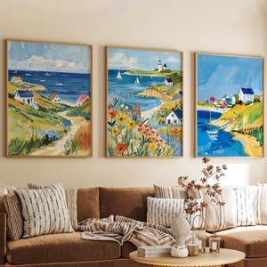 May include: Three framed landscape paintings showcase coastal scenes with houses, sailboats, and a lighthouse. The artwork uses vibrant colours, including blues, greens, yellows, and reds, with a focus on the sea and sky. The paintings are displayed above a brown sofa with striped cushions.