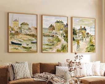 May include: Three framed watercolor paintings depicting coastal scenes. The artwork features buildings, boats, and people, rendered in soft greens, yellows, and blues. The paintings are displayed above a brown sofa with striped pillows, creating a cohesive and inviting aesthetic.