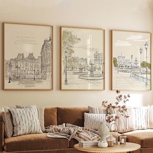 Op de afbeelding: Drie ingelijste kunstprints met Europese stadsgezichten in een schetsstijl. Het kunstwerk toont gebouwen, fonteinen en straatlantaarns in zwart-wit met groene en beige accenten. De frames zijn licht van kleur.