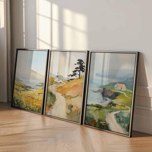 May include: Three framed landscape paintings. Each features a coastal scene with a winding road, a house, and a tree. The artwork has a soft, watercolor style with muted colors of green, yellow, and blue. The frames are black.