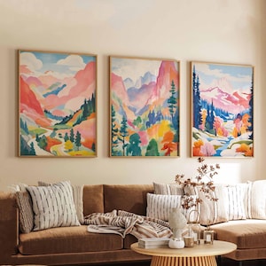 May include: Three framed watercolor landscape paintings. Each features a mountain scene with vibrant colors, including pink, blue, green, and yellow. The paintings are displayed above a brown sofa with striped pillows and a wooden coffee table.