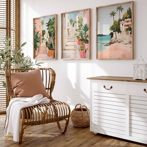 May include: A wicker chair with a brown cushion and a white throw blanket sits in a room with three framed paintings of coastal scenes. A white cabinet with two drawers and a wooden top is in the background.