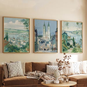 May include: Three framed landscape paintings with a vintage aesthetic. The artwork depicts European cityscapes and coastal scenes in shades of blue, green, and beige. The paintings are displayed above a brown sofa with striped pillows.
