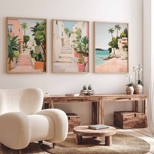 May include: Three framed illustrations of colorful scenes with buildings, palm trees, and stairs. The illustrations are hung on a white wall above a wooden table and a white armchair.