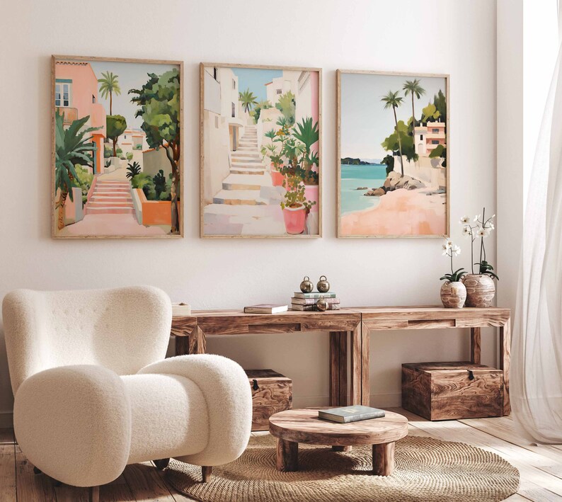 May include: Three framed illustrations of colorful scenes with buildings, palm trees, and stairs. The illustrations are hung on a white wall above a wooden table and a white armchair.