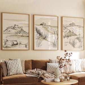 May include: A set of three framed wall art prints featuring line drawings of coastal landscapes with castles. Each print displays a beige background with black ink sketches of dunes, grasses, and stone structures situated near the shoreline.