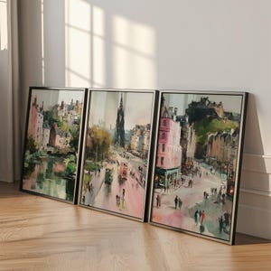May include: Three framed watercolor prints of a city street scene. The artwork features buildings in shades of pink, brown, and green, with people and vehicles on the street. The frames are black with silver inner edges.