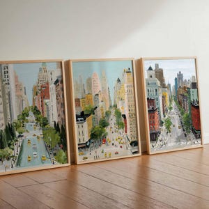 May include: Three framed art prints depicting a city street scene with tall buildings, trees, and yellow taxis. The artwork is in a watercolor style, with a light blue sky and a wooden frame.