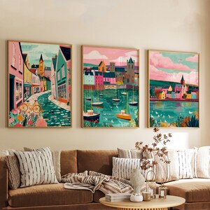 May include: Three framed art prints depicting colorful coastal scenes. The artwork features buildings, boats, and water in vibrant hues of pink, teal, and yellow. The prints are displayed above a brown sofa with striped pillows.