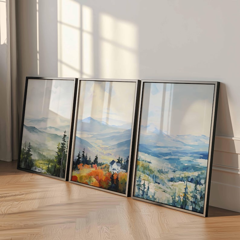 May include: Three framed landscape art prints. Each print features a watercolor painting of a mountain scene with trees and a house. The frames are black with a silver border, and the artwork has a soft, muted color palette.