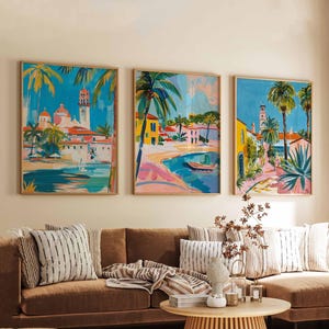 May include: Three framed art prints showcasing coastal scenes with palm trees and buildings. The artwork uses a vibrant palette of blues, yellows, pinks, and greens. The prints are displayed above a brown sofa with decorative cushions.