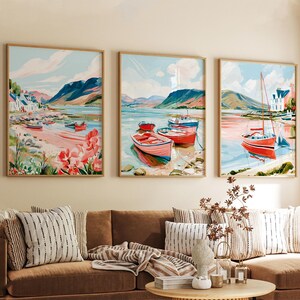 May include: A set of three framed wall art prints depicting coastal landscapes with mountains, calm water, and red boats. The paintings feature a light blue sky, pinkish shorelines, and green hills, all enclosed in thin, light-colored wooden frames.