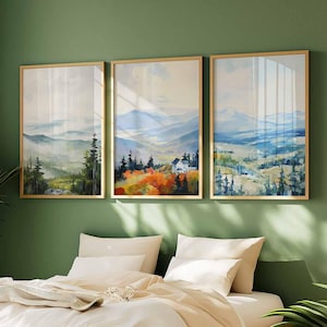 May include: Three framed landscape art prints. Each print features a watercolor painting of a mountain scene with trees and a house. The frames are gold-colored, and the artwork is displayed on a green wall above a bed with white bedding.