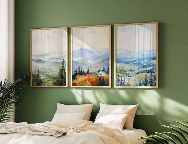 May include: Three framed landscape art prints. Each print features a watercolor painting of a mountain scene with trees and a house. The frames are gold-colored, and the artwork is displayed on a green wall above a bed with white bedding.