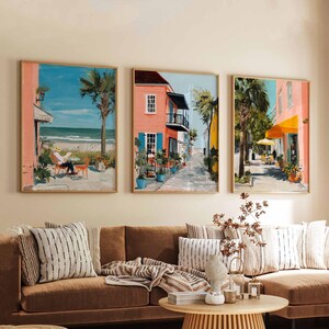 Peut inclure: Trois impressions d'art encadrées représentant des scènes côtières. Les peintures présentent des bâtiments colorés, des palmiers et des vues sur la plage. L'œuvre est exposée au-dessus d'un canapé marron avec des coussins à motifs et une table basse en bois.