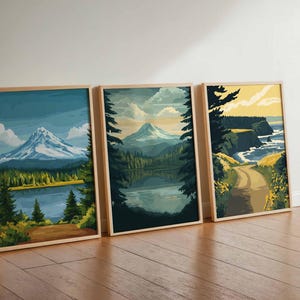 May include: Three framed landscape art prints. The prints depict mountain and lake scenes with a color palette of blues, greens, and yellows. The frames are made of light-colored wood. The art is leaning against a white wall on a wooden floor.