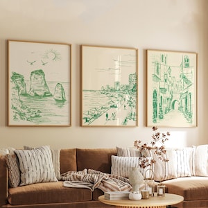 Peut inclure: Trois impressions d'art encadrées en vert et blanc. Les impressions représentent des scènes côtières, notamment des formations rocheuses, une promenade en bord de mer et une scène de rue avec des arches. Les impressions sont affichées au-dessus d'un canapé marron avec des coussins rayés.