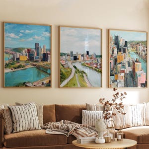 Op de afbeelding: Drie ingelijste stadsgezichtschilderijen met houten frames hangen boven een bruine bank. De schilderijen tonen een stad met hoge gebouwen, bruggen en een rivier. Het kunstwerk heeft een schilderachtige stijl met een blauw, groen en geel kleurenpalet.