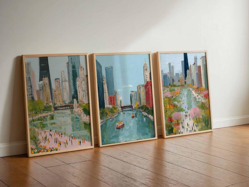 May include: Three framed cityscape paintings. Each features a river scene with buildings, bridges, and people. The artwork has a light color palette with blue skies, and the frames are light wood.