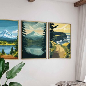 May include: Three framed landscape art prints. The prints depict mountain and lake scenes with a road leading to the coast. The color palette includes blues, greens, and yellows, with gold frames. The art is displayed on a white wall.