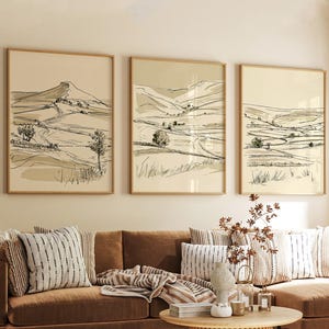 May include: A set of three framed wall art prints featuring black line drawings of rolling hills, fields, and trees on a cream-coloured background. Each rectangular print is encased in a thin, light-toned wooden frame, displaying a landscape.