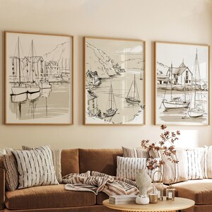 Op de afbeelding: Een set van drie ingelijste wandkunstwerken met zwart-witte lijntekeningen van zeilboten in een haven met kustgebouwen. De kunstwerken hebben een neutrale beige achtergrond en hangen boven een bruine bank met kussens met een patroon.